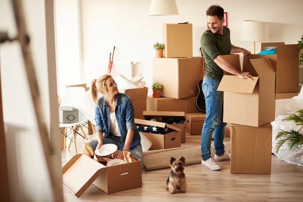 Couple unpacking cardboard boxes in their new living room as a small dog looks on.