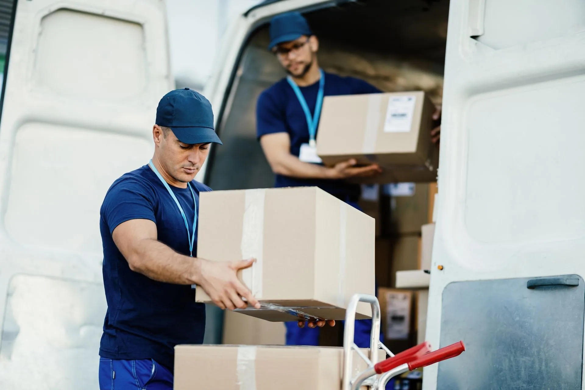 Two movers in blue caps unloading cardboard boxes from a white delivery van.