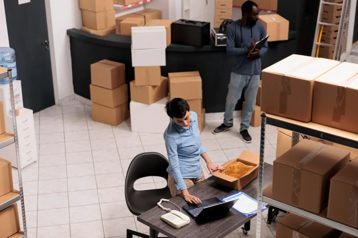 Two workers checking stacked boxes and inventory on a laptop in an office storeroom.